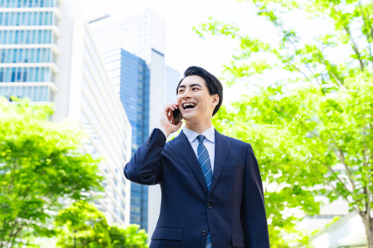 新緑の街でスマホで通話する男性ビジネスパーソン