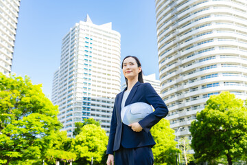 新緑の街の前にヘルメットを持って立つ女性ビジネスパーソン