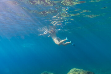 Fototapeta premium Underwater shot of a young woman in a bikini and fins gracefully swimming in the clear blue sea with sun rays.