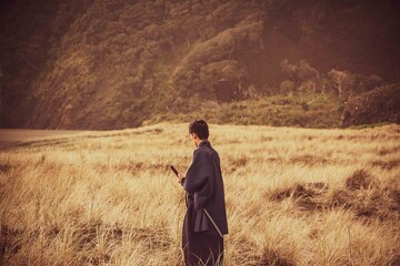 A samurai in black is running via meadow and forest nearby ocean in New Zealand.