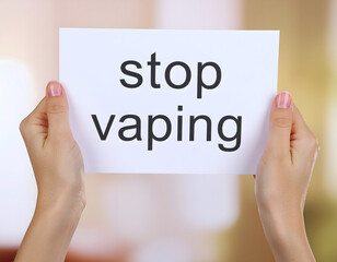 Two light-skinned hands gently hold a white rectangular paper displaying the text "stop vaping" in a clear, bold black font. The background is softly blurred