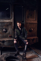 Handsome individual sits on a worn tire, surrounded by a shattered train, embodying resilience.