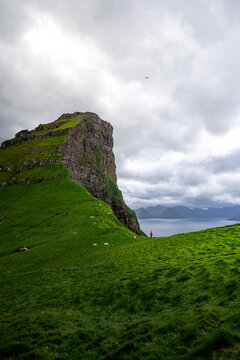 View of a dramatic cliff face rises from a vibrant green hillside, contrasting against the muted blues of the distant sea, Kallur, Nor&Atilde;&deg;oya, Faroe Islands.
