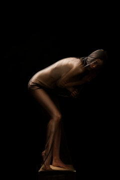 A woman draped in sheer fabric bends forward, her face obscured by the material. She stands on a platform against a dark background, creating a dramatic and artistic pose.