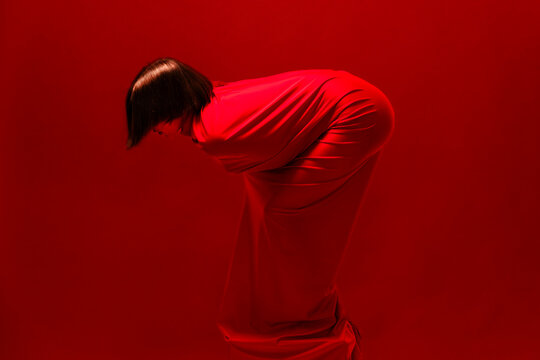 A person in a flowing red garment bends forward against a vibrant red background. The figure's posture conveys a sense of introspection, submission, or artistic expression.