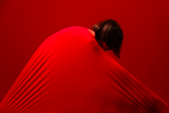 A person is enveloped in a vibrant red fabric, their head and dark hair visible, looking down. The stretched cloth creates dynamic lines against the matching red background. An abstract image.