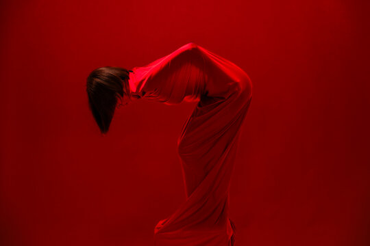 A person is fully covered by a flowing red fabric, bending deeply with their head bowed. Dark hair cascades downwards. The monochromatic red setting emphasizes the dramatic and mysterious pose.