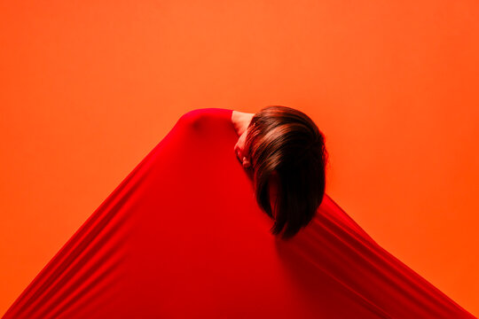 A woman's head with dark hair is seen pushing through a vibrant red fabric. The fabric stretches tautly against a bright orange background, creating an abstract and striking visual.