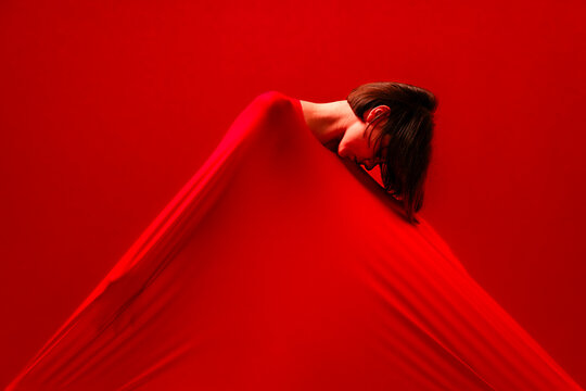 A woman's head and shoulders are visible, emerging from a large red fabric. The red cloth drapes around her, creating an abstract and artistic composition against a red background.