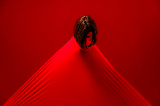 A woman's head emerges from a taut red fabric, forming a striking triangular shape against a red background. The fabric stretches downwards, creating strong lines and shadows.