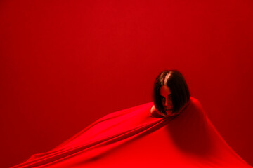 A young woman with dark hair emerges from vibrant red fabric against a matching red background. She looks directly at the camera with an intense gaze. This abstract image conveys emotion and mystery.