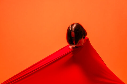 A person with dark hair emerges from a large red fabric, creating a striking visual against an orange background. This artistic shot emphasizes color and form.