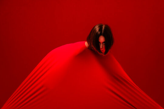 A woman's head emerges from a large, stretched red fabric. The vibrant red material creates a striking, abstract composition against a matching red background.