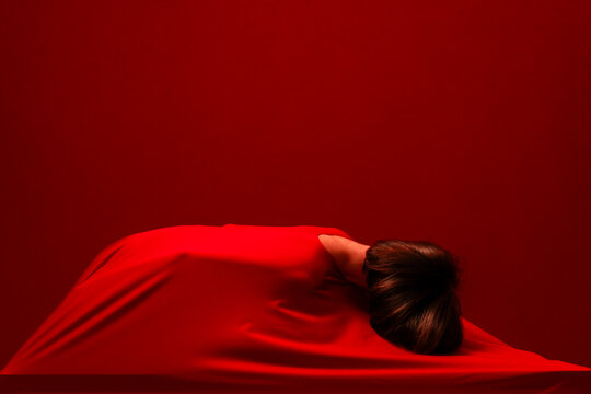 A woman is covered by a vibrant red fabric, creating an abstract and mysterious form. Her head is visible from behind, resting on the draped cloth against a matching red background.