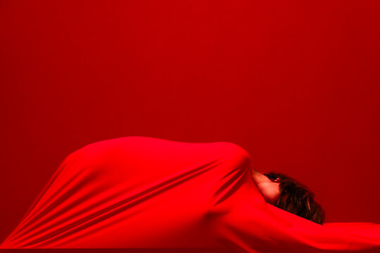 A person's head and hair emerge from vibrant red fabric, which drapes over their body. The red cloth creates soft folds against a matching red background, forming an abstract composition.