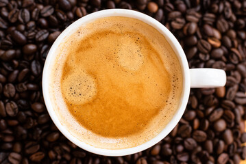 Top view of a cup of black coffee with crema, surrounded by roasted coffee beans on a dark background.