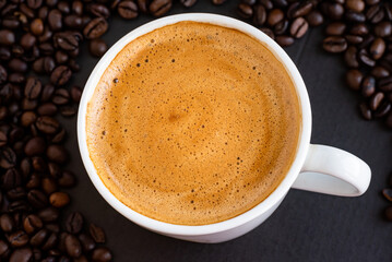Top view of a cup of black coffee with crema, surrounded by roasted coffee beans on a dark background.