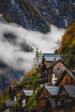 View of quaint wooden houses nestled on a hillside shrouded in mist, contrasting with the rugged, forested mountains, Hallstatt, Austria.
