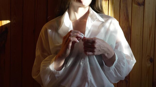 Woman unbuttoning shirt, young female unbuttons sheer white blouse, revealing bra in intimate morning light