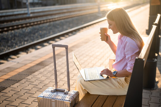 Elegant businesswoman using laptop and drinking coffee while sitting on a bench with luggage at the sunny railway station and waiting arrival of train.