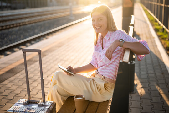 Portrait of elegant businesswoman using smartphone while sitting on bench at the railway station with suitcase and waiting arrival of train. - Powered by Adobe