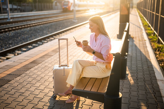 Elegant businesswoman using smartphone and drinking coffee while sitting on a bench with suitcase at the railway station and waiting arrival of train.