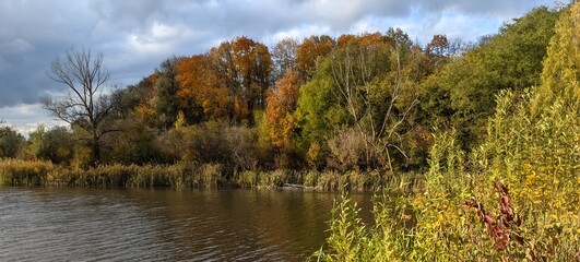Golden autumn. Trees dressed in crimson and gold robes are reflected in the mirror-like surface of the river. The sky is covered with light clouds. Sunbeams break through them