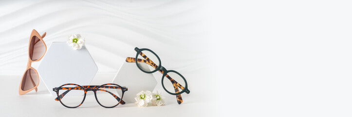 Optic store advertisement banner. New trendy eyewear collection on a white Background with flowers. Clear Eyeglasses and sunglasses on podiums. Minimal style. Copy space. Summer sale special offer