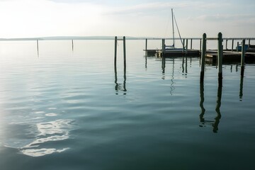 Obraz premium A serene scene of a quiet lake with a small pier extending into the water, featuring a sailboat docked at the end.