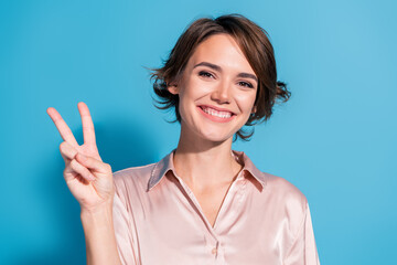 Fototapeta premium Charming young woman making peace sign with a big smile in elegant satin blouse against a vibrant blue background