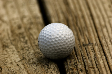Golf ball rests on weathered wooden bench with dimpled surface centered in frame. Wood grain and natural imperfections highlight outdoor stillness.