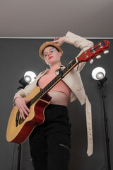 Portrait of confident female guitarist holding guitar, looking at camera. With one hand she holds brim of her straw hat, her stage outfit complemented by leather jacket and jeans. Low angle view