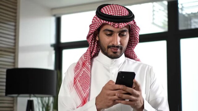 Young Arab businessman in traditional Saudi thobe and keffiyeh using a smartphone in a modern office, symbolizing the integration of culture and contemporary technology.