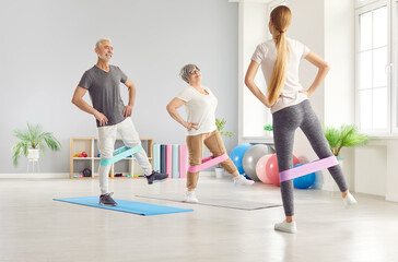 Senior couple working out together with a coach, using resistance bands for stretching exercises in the gym. Fitness routine focuses on flexibility and teamwork during the training session.