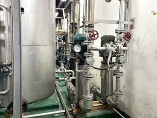 Close-up of industrial stainless steel pipelines, valves, and gauges connected to a large tank inside a modern industrial facility or boiler room, showcasing engineering and manufacturing technology.