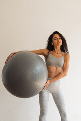 Fototapeta premium Healthy middle-aged woman in sportswear confidently holding a large exercise ball while standing barefoot on a yoga mat in a minimalist home studio, symbolizing vitality, fitness, and positive lifesty