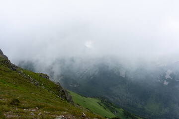 Steep mountain slope with rocky outcrops overlooking fog-filled valley below on overcast day creating dramatic alpine scenery. Concept of rugged terrain exploration and challenging hiking conditions.