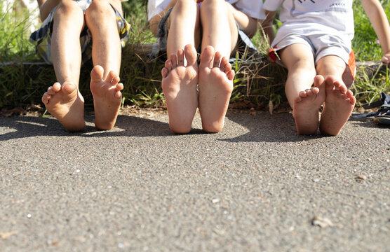 Cute bare feet of three children on asphalt on a sunny day. Children, brothers, sisters or friends took off their shoes and are relaxing sitting on the side of the road. Summer fun, freedom.
