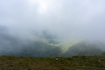 Dense cloud cover revealing glimpses of lush green mountain valleys through swirling mist from high elevation viewpoint. Concept of dramatic mountain meteorology and elevated perspective landscapes.