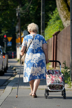 An elderly woman with a walking cane shopping trolley walks on a city sidewalk. This authentic rear view photo illustrates an active senior lifestyle, independence, mobility, daily life of a retiree