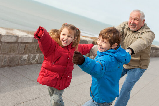 two cute kids, little boy and girl, playing with their grandfather outdoors pulling his hands, laughing and  running on seaside promenade on cold sunny  day.  - Powered by Adobe