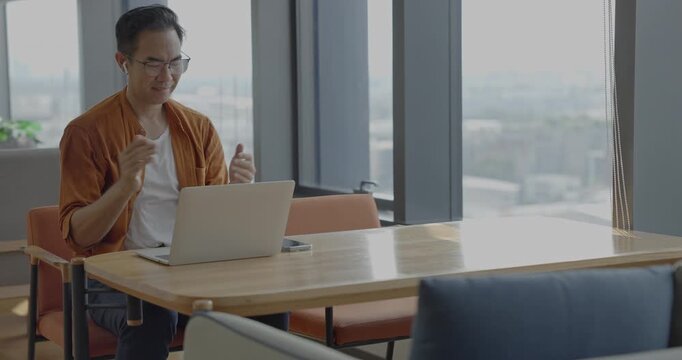 Senior Asian businessman wearing headset celebrates success during online meeting in modern co-working space. Remote work and business communication concept