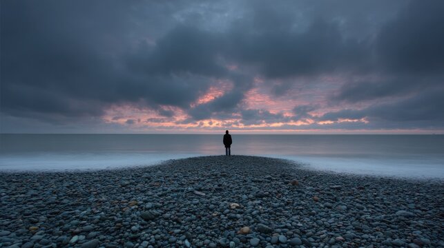 Solitary Figure Standing on Rocky Shore at Sunset with Dramatic Sky - Powered by Adobe