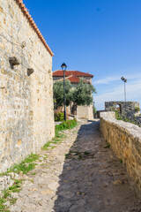  Narrow stone walkway in Ulcinj’s Old Town, Montenegro, leading past ancient walls to a seaside lookout. The Adriatic Sea glistens under a clear blue sky beside historic fortress structures.