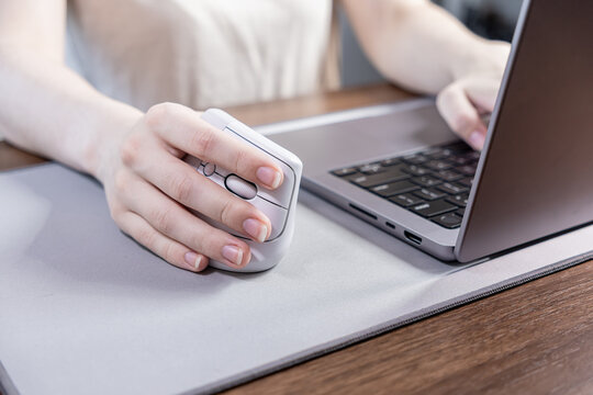 A person's hand uses an ergonomic gray vertical mouse, promoting wrist comfort and efficiency while working on a modern laptop at a desk