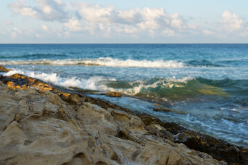 Rocky shoreline with gentle waves crashing under a partly cloudy sky, showcasing the natural textures of rocks and the calm blue ocean horizon. Sea. Water. Summer. Rocks