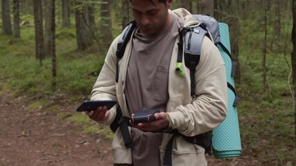 Tilt up shot of young male intercultural tourist connecting mobile phone and GPS navigator while exploring trails in lush forest - Powered by Adobe
