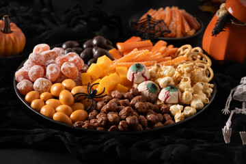 Halloween sweet charcuterie board with pumpkin, popcorn, orange chocolate candy, spider, crackers on black background. Scary decoration. Festive food for kids party. Close up.
