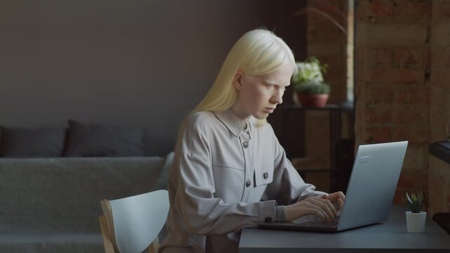 Androgynous young woman with albinism sitting at desk in loft living room at home texting something on laptop