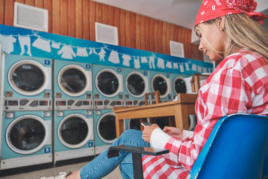 Young woman using smartphone while waiting for laundry, sitting on chair in laundromat - Powered by Adobe